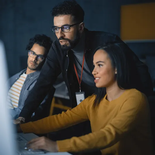 Three software developers talking about programming issue leaned over desk looking at computer monitor