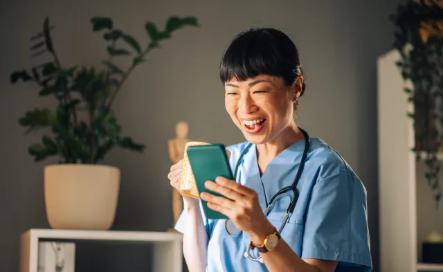 Asian female nurse in blue scrubs laughs heartily while looking at her mobile phone, holding a mug of coffee. The background features potted plants, bookshelves, and a small wooden figure.