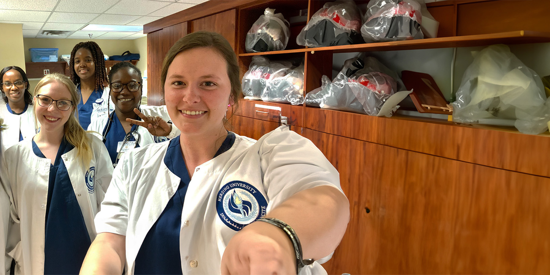 Five Herzing nursing students in white coats and blue scrubs take a selfie in a lab, with medical equipment wrapped in bags right beside them.