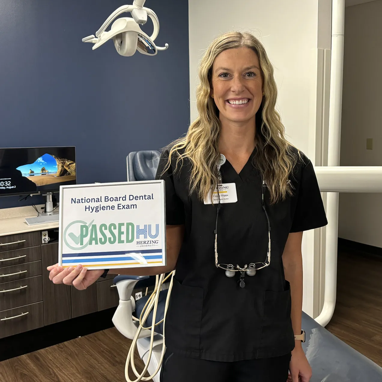 A female dental hygiene student in black scrubs smiles while holding a ‘Passed National Board Dental Hygiene Exam’ certificate in a dental clinic setting, with a Herzing University ID badge visible.