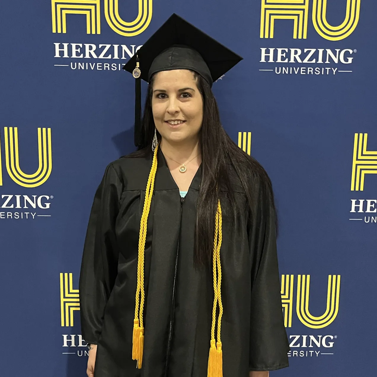 A Herzing graduate in a black graduation gown and cap stands smiling in front of a Herzing University banner, wearing two yellow honor cords around her neck.