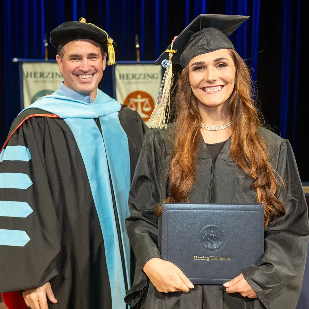 A proud graduate in a black cap and gown holding her diploma while standing next to a professor in academic regalia, with Herzing University banners in the background during a graduation ceremony.