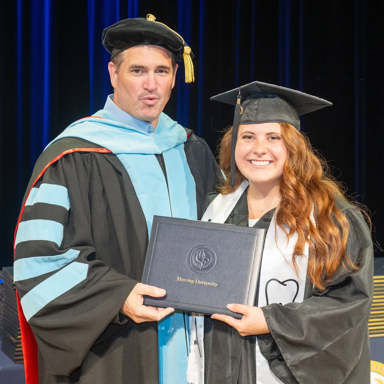A Herzing University graduate receiving her diploma alongside a faculty member during a commencement ceremony, symbolizing academic excellence.