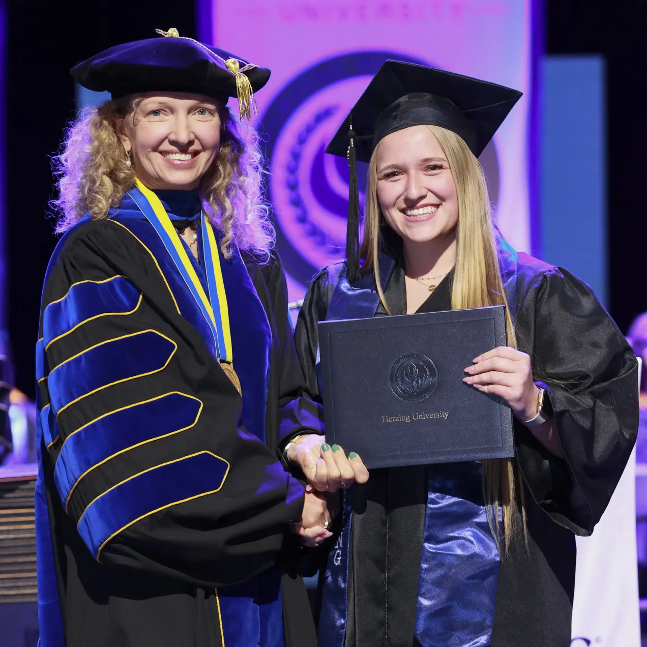 A Herzing University graduate smiling as she receives her diploma on stage, alongside a faculty member in ceremonial regalia during commencement.