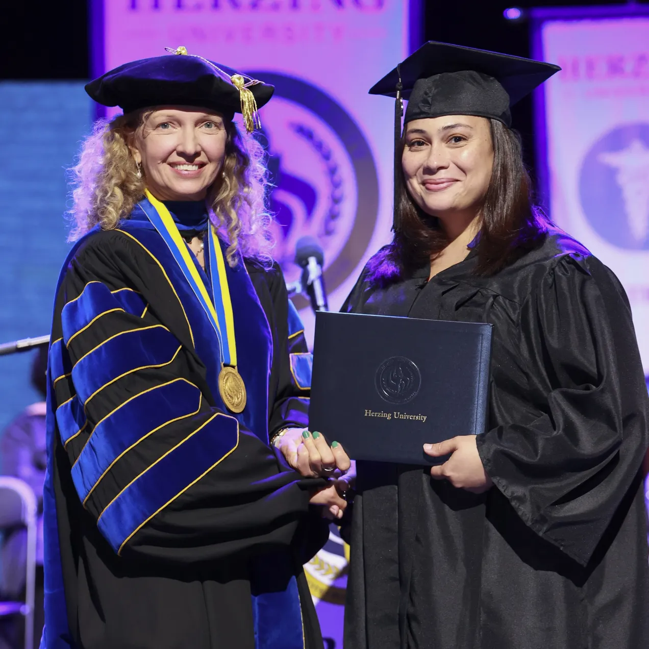 A Herzing University graduate receiving her diploma on stage, accompanied by a faculty member in ceremonial regalia.