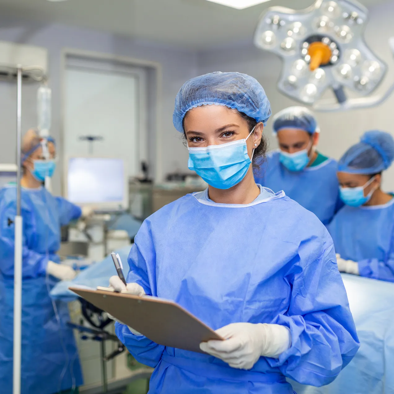 Surgical technology student in scrubs and a mask holding a clipboard in an operating room setting with other students practicing procedures in the background.