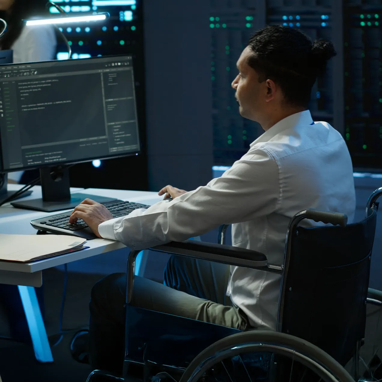 A focused cybersecurity professional working in a wheelchair at a desk in a high-tech server room, analyzing code on a large monitor, highlighting inclusivity and the practical skills emphasized in Herzing University’s graduate-level cybersecurity program.