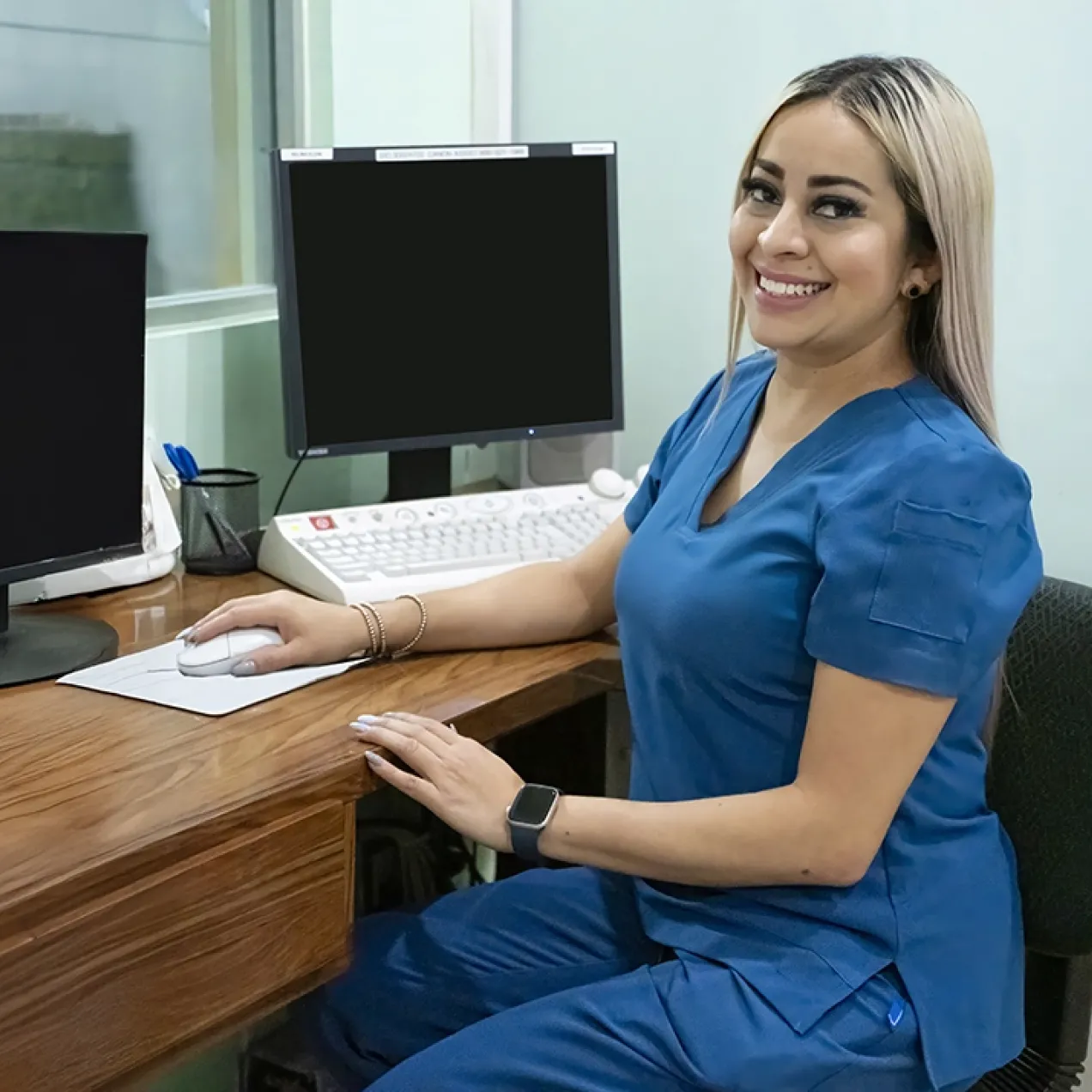 Medical office professional in blue scrubs seated at a desk with dual monitors, showcasing a career outcome of the Medical Office Administration program.