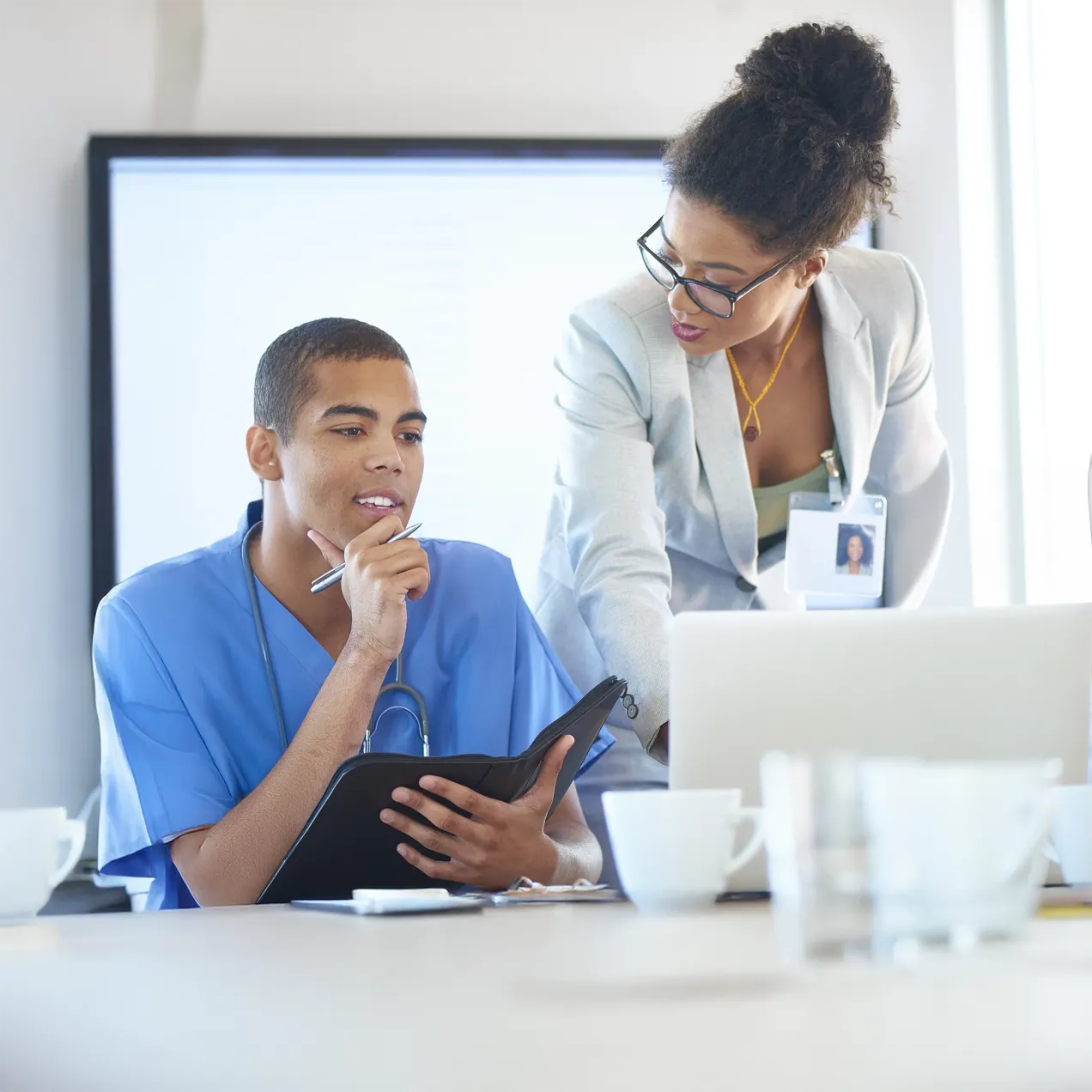 Healthcare professional in scrubs reviewing documents while collaborating with a colleague in a business suit in a meeting room.