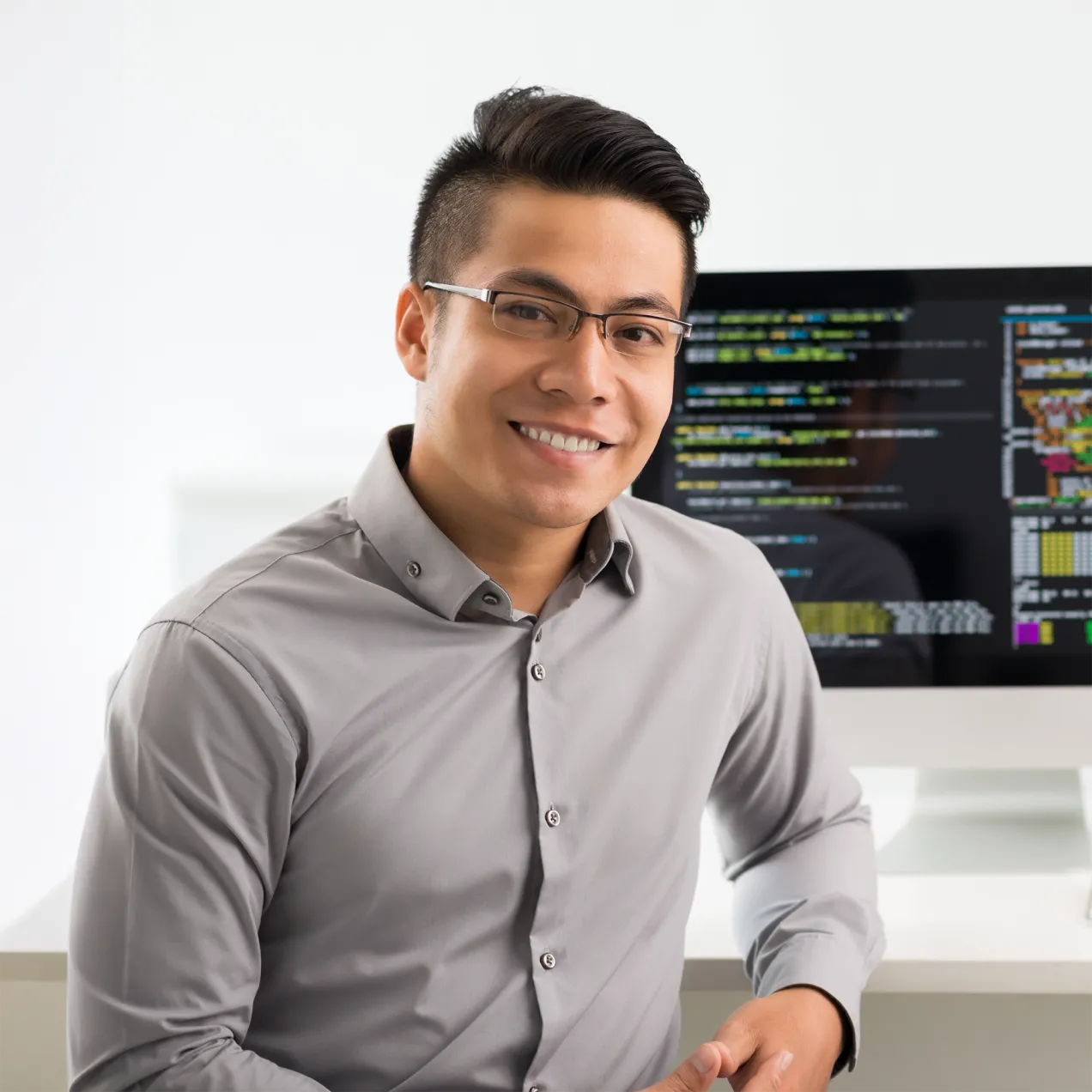 A smiling cybersecurity professional in a modern office setting, seated in front of a computer displaying code, representing the knowledge and skills developed in Herzing University’s cybersecurity program.
