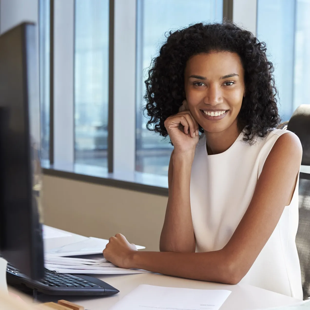 Confident businesswoman sitting at her desk in a bright office, smiling at the camera with a computer monitor and paperwork in the foreground.