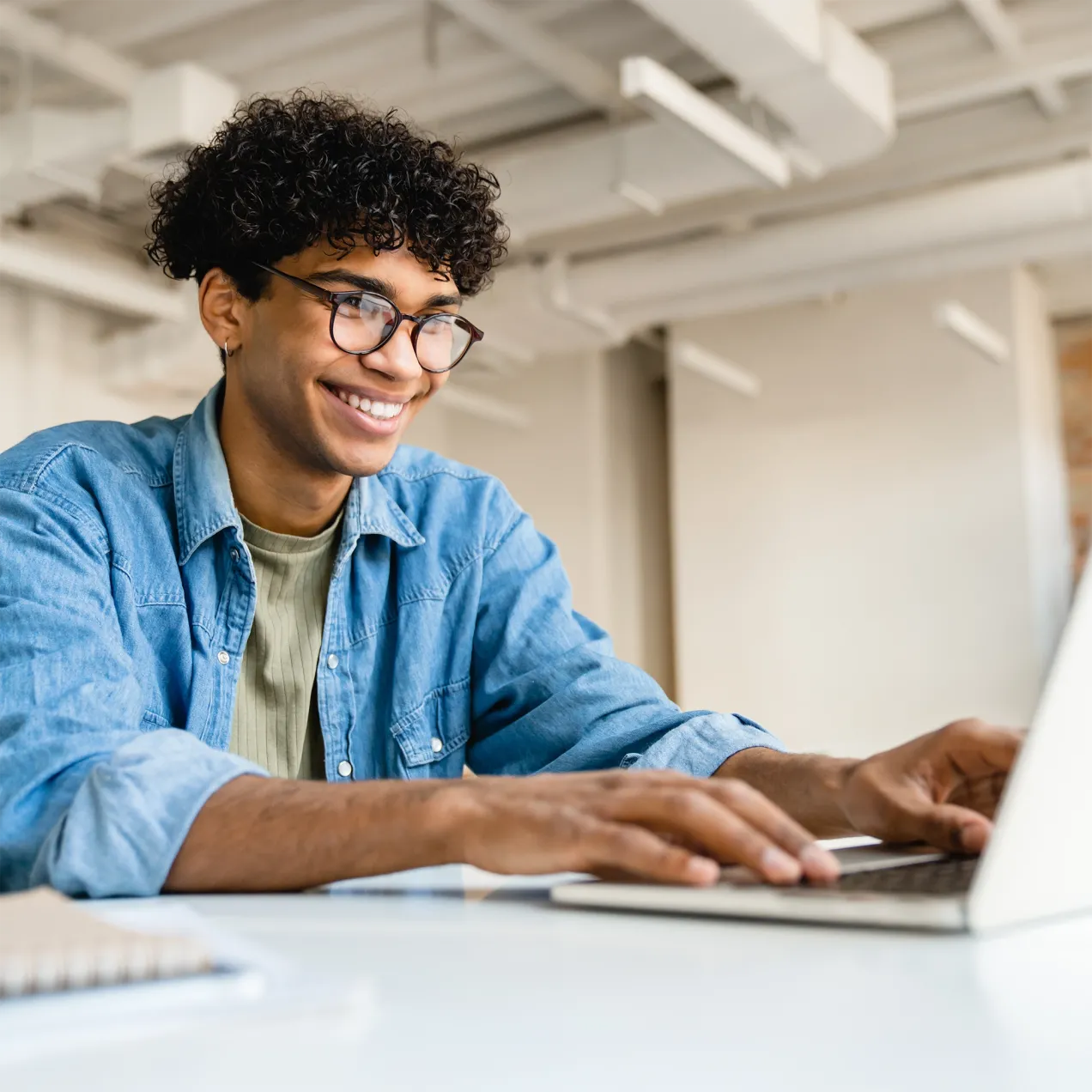 A Herzing University information technology student in a denim shirt and glasses smiles while working on a laptop in a modern, well-lit workspace.