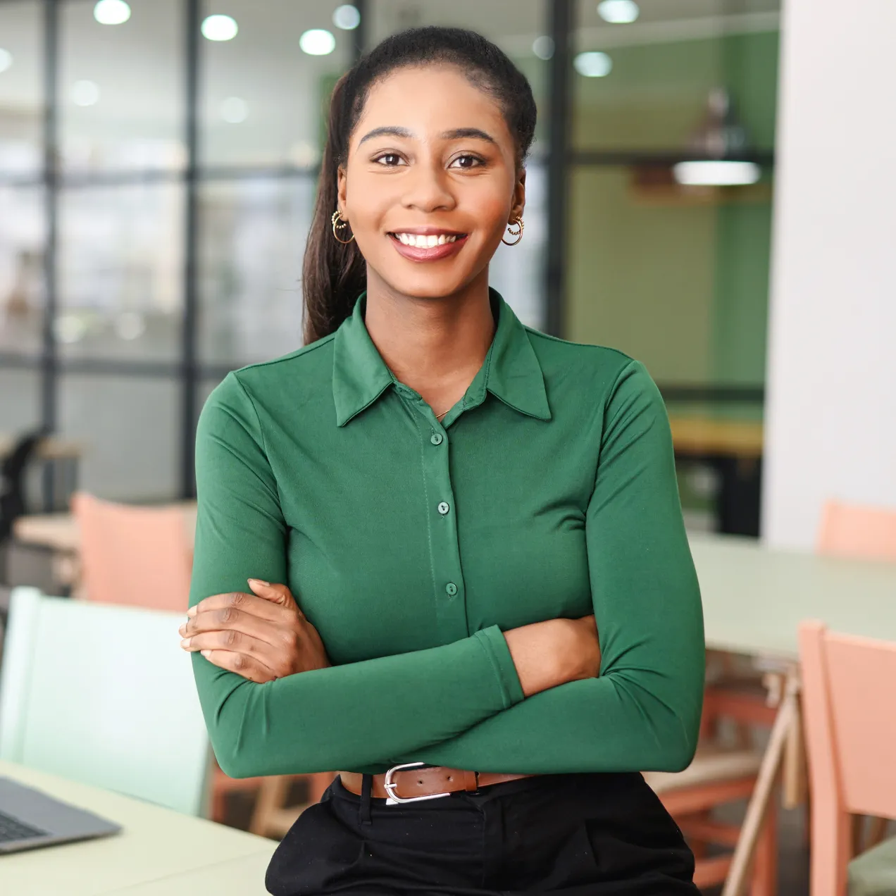 A professional woman in a green blouse sits at a modern office table, smiling with arms crossed, ready for career success.