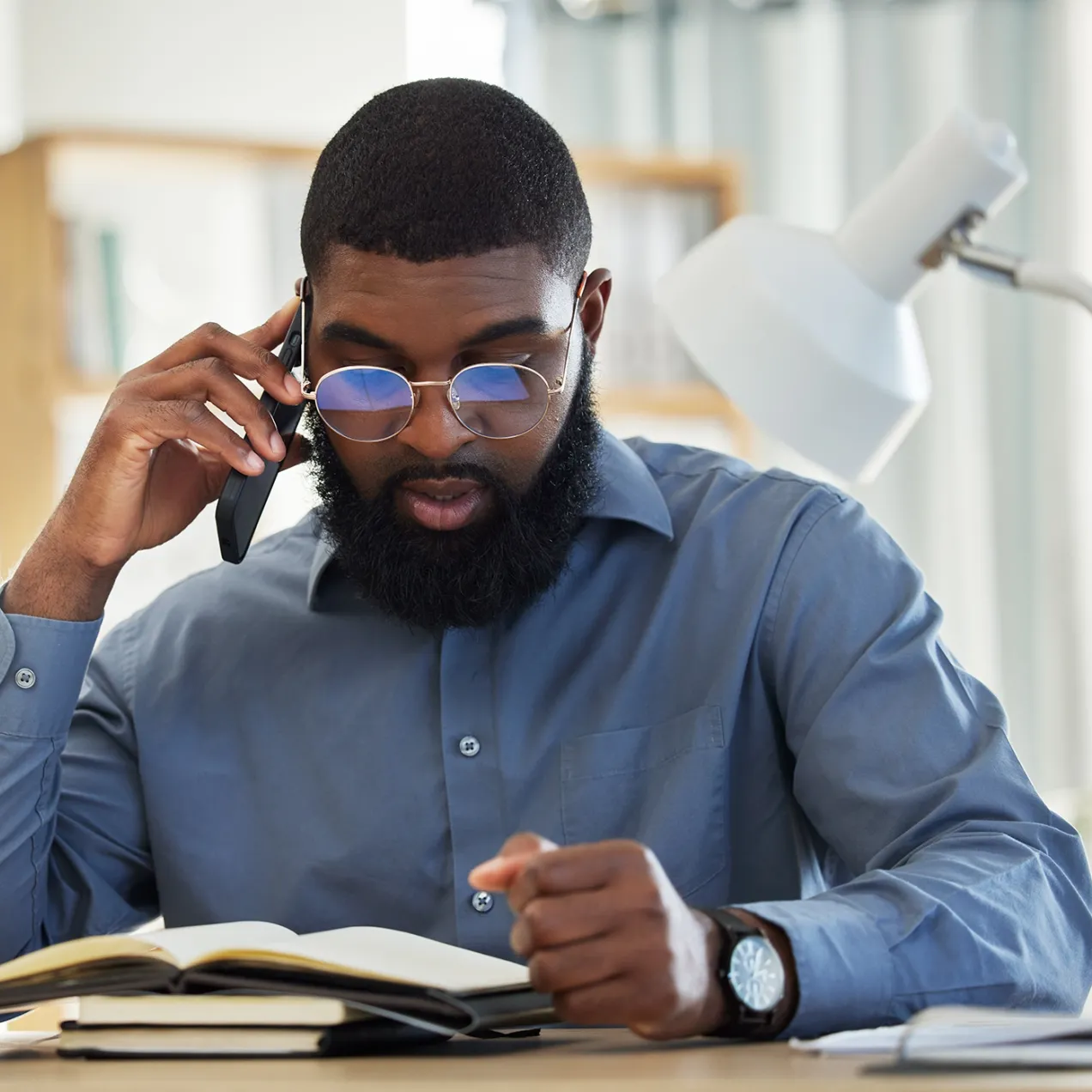 A criminal justice professional in a blue dress shirt and glasses reads a legal book while speaking on the phone, with a scale of justice and a desk lamp in the background.