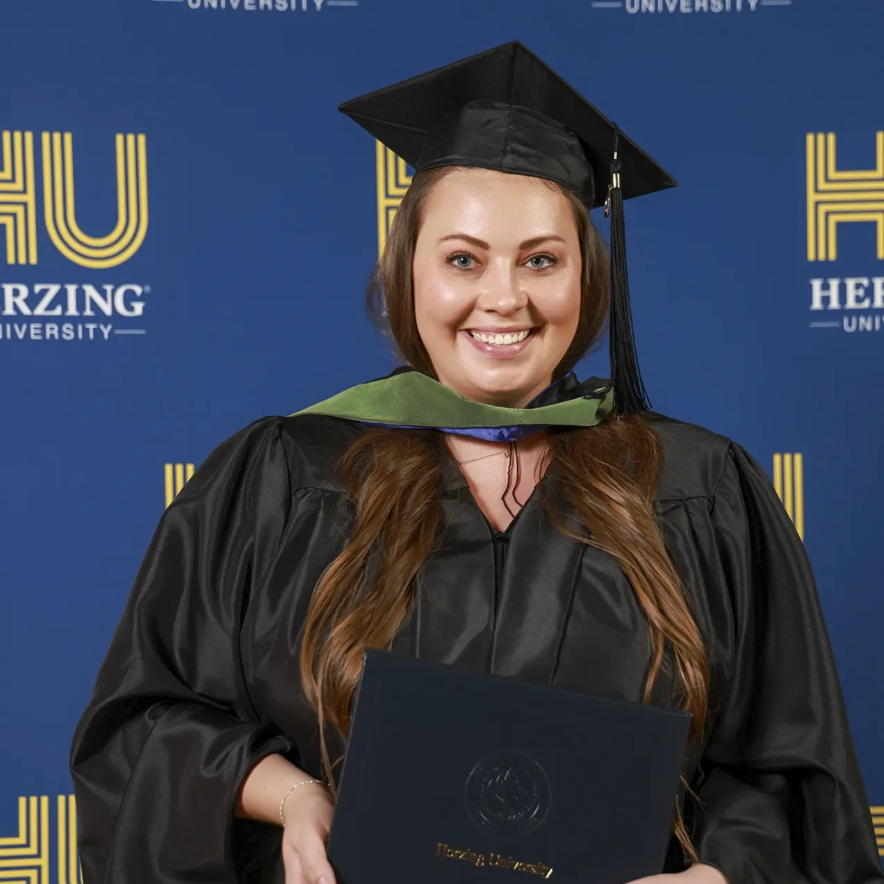 Proud Herzing University graduate in cap and gown holding diploma in front of official university backdrop.