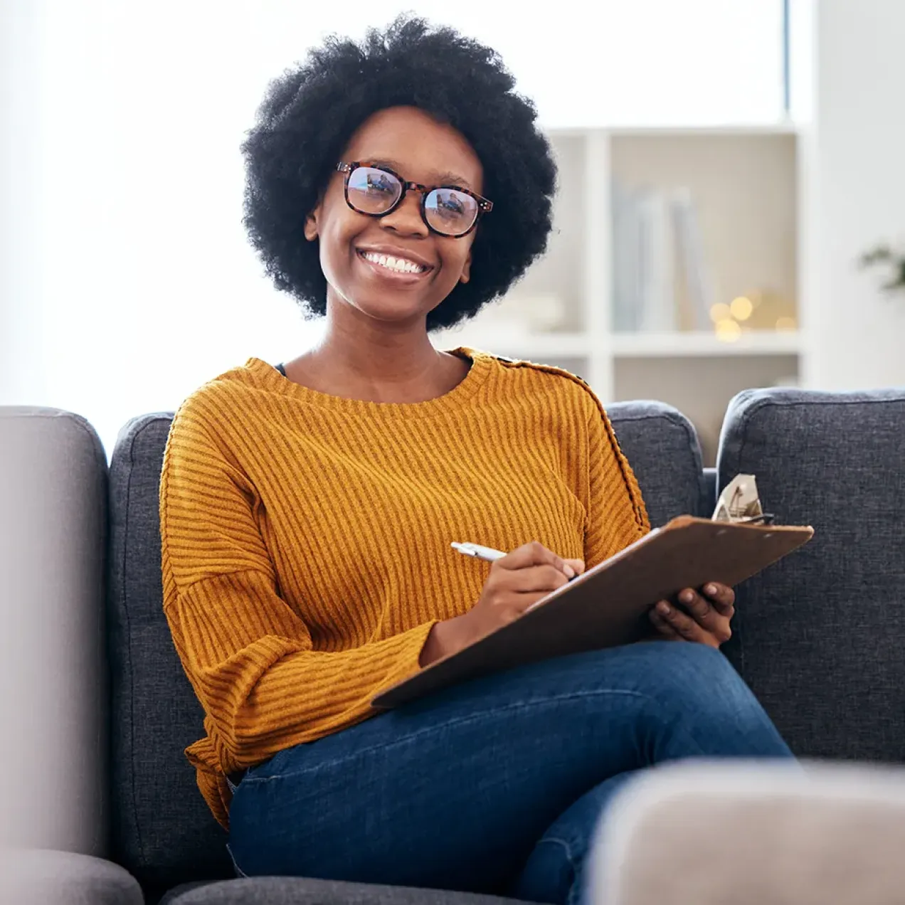 Smiling counselor with natural hair and glasses sitting on a couch, holding a clipboard and pen during a therapy session.