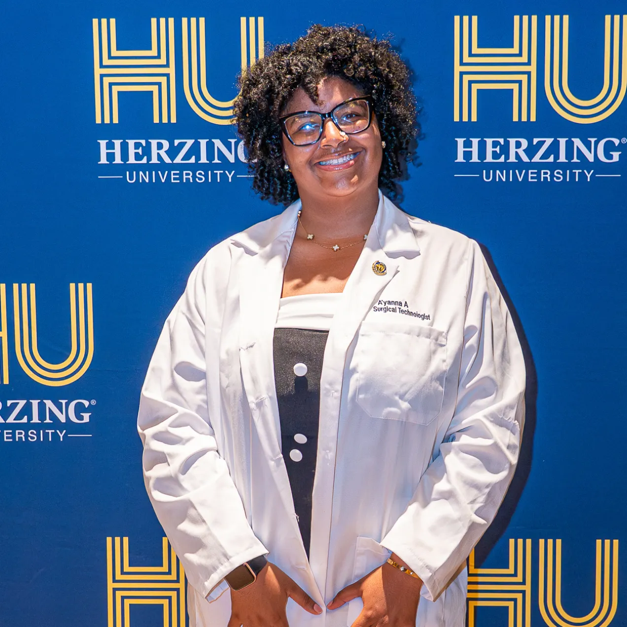 Herzing University Surgical Technology student wearing a white lab coat and glasses smiles in front of a blue Herzing University backdrop.