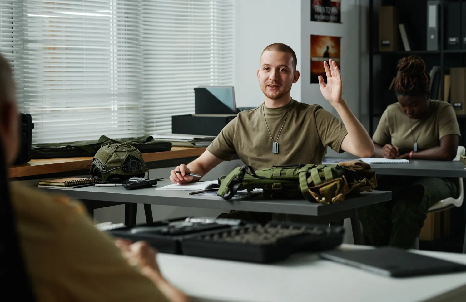 Young male student raising hand and looking at teacher at lesson while asking him question after lecture about military training