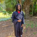 Joe Johnson in a grove of palm trees in a graduation gown holding a grad cap