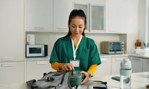 Healthcare professional packing a medical bag while standing in a contemporary kitchen setting with various kitchen appliances visible in background