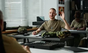 Young male student raising hand and looking at teacher at lesson while asking him question after lecture about military training