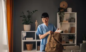 Asian dedicated nurse in blue scrubs is seen preparing medical tools and organizing documents into a brown backpack in a well-lit, modern room with shelves and assorted decor.