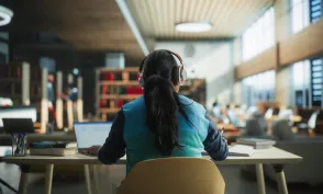 Female Student Wearing Headphones while Working on University Homework in a Public Library. Woman Sitting Behind a Desk, Using Laptop Computer and Writing Down Notes in Notebook. Footage from the Back