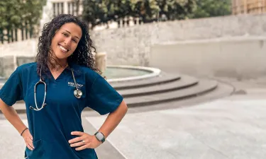 Herzing University online nursing student in blue scrubs and hands on hips smiling outside in courtyard