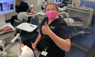 A dental hygiene student giving a thumbs up while performing a dental procedure. The student is wearing a pink mask and protective glasses, with a dental monitor showing images in the background. A patient is seated in the dental chair, receiving treatment.