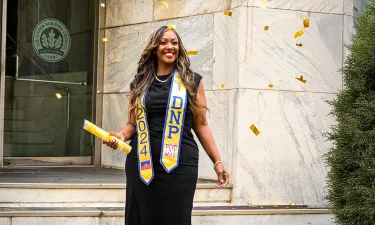 Graduating student celebrating with confetti, wearing a black dress and DNP 2024 sash, holding a diploma outside a building.