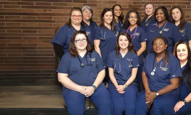 Group of LPN to RN students wearing blue scrubs smiling for picture in front of brick wall