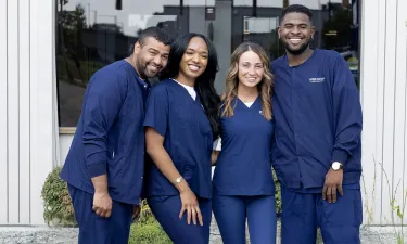 Wide-angle group photo of Herzing University medical assisting students smiling in navy scrubs outside a campus building, emphasizing teamwork and inclusivity.