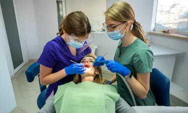 Two dental professionals in protective gear and masks performing a dental procedure on a patient in a modern dental office, showcasing teamwork and hygiene practices.