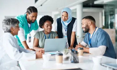 Diverse healthcare professionals collaborating around a laptop during a meeting, demonstrating teamwork and leadership in healthcare administration.