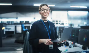 A smiling cybersecurity professional holding a laptop in a high-tech office environment, with multiple monitors displaying analytical data, symbolizing the cutting-edge expertise gained in Herzing University’s cybersecurity program.