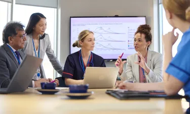 Healthcare administrators and professionals collaborating in a meeting room with a presentation displayed on a screen, discussing data and strategies.