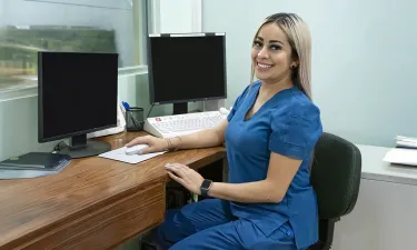 Medical office professional in blue scrubs seated at a desk with dual monitors, showcasing a career outcome of the Medical Office Administration program.