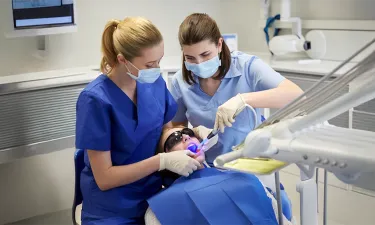 Dental assisting students wearing scrubs and masks working together on a patient in a clinical setting, demonstrating hands-on training in a dental assisting diploma program.