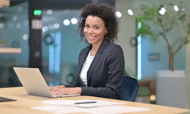 A professional woman sits at a desk with a laptop, smiling confidently, representing career success with a business management degree.