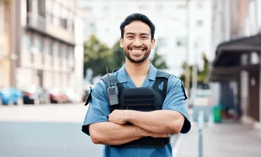 A Herzing University criminal justice student in a police uniform and tactical vest stands confidently with arms crossed, smiling in an urban setting.