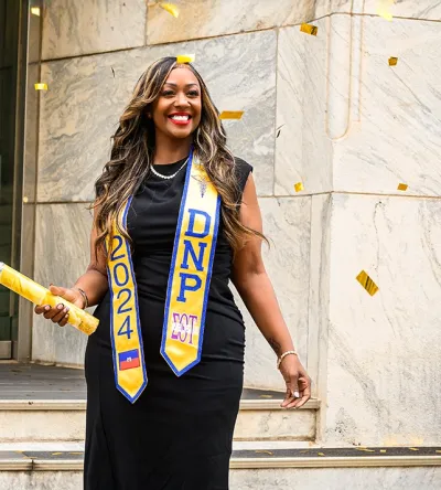 Graduating student celebrating with confetti, wearing a black dress and DNP 2024 sash, holding a diploma outside a building.