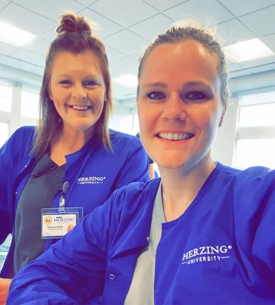 Two Herzing University students in blue scrub jackets pose for a selfie, smiling and wearing ID badges, with a bright office background.