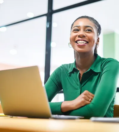 A smiling woman in a green shirt sits at a desk with a laptop in front of her, looking upbeat and engaged. She appears to be in a modern workspace with bright lighting.