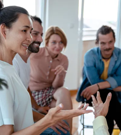 A diverse group of people is seated in a circle in a group therapy or support session. A woman in white is speaking, and others, including a man and a woman in a red shirt, listen and engage with her. The room has bright windows and a warm atmosphere.