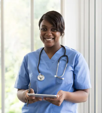 Smiling medical assistant in blue scrubs with a stethoscope, holding a tablet indoors.