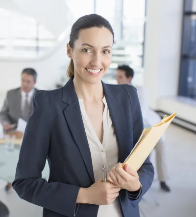 Healthcare administrator smiling in a professional office setting while holding a folder, with a team discussing in the background.