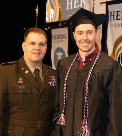 Herzing University graduate in cap and gown posing with a military service member in uniform during a graduation ceremony.