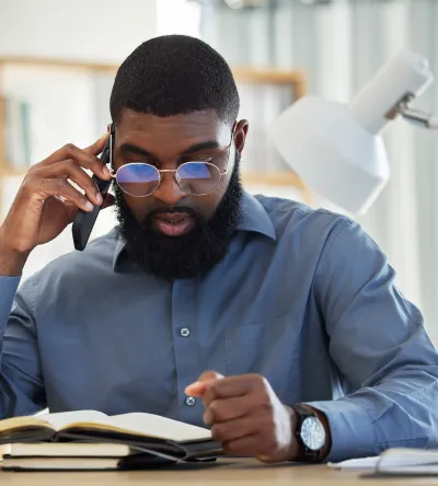 A criminal justice professional in a blue dress shirt and glasses reads a legal book while speaking on the phone, with a scale of justice and a desk lamp in the background.