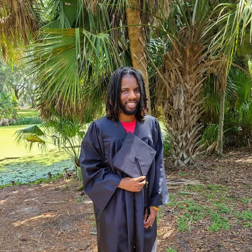 Joe Johnson in a grove of palm trees in a graduation gown holding a grad cap