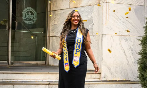 DNP banner Graduating student celebrating with confetti, wearing a black dress and DNP 2024 sash, holding a diploma outside a building.